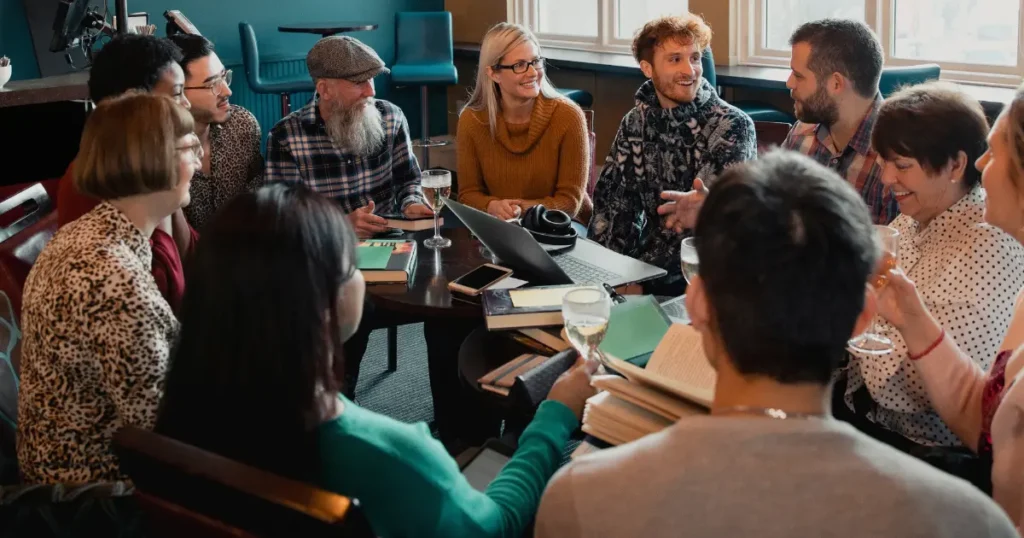Fotografia calda e luminosa con un focus morbido. La scena mostra un gruppo eterogeneo di persone (età e generi diversi) sedute in cerchio in un ambiente accogliente, come una biblioteca comunitaria o una sala con piante e luce naturale. Stanno collaborando a qualcosa di creativo: alcuni sfogliano libri, altri scrivono su un grande foglio di carta comune al centro. Le loro espressioni sono concentrate ma serene. L'immagine non deve mostrare volti specifici in primo piano, ma catturare il senso di unione, collaborazione e crescita condivisa. Lo stile è autentico e umano, evocando un forte senso di community e di scopo comune.