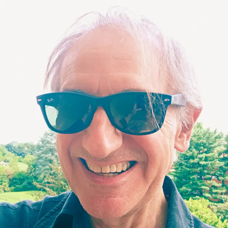 Primo piano di Maurizio Bizzozero, autore di Poliedricamente Edizioni, che indossa occhiali da soli neriè sorridente e sullo sfondo ci sono un prato e degli alberi.