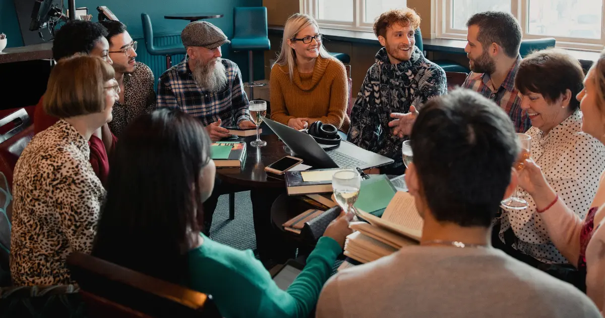 Fotografia calda e luminosa con un focus morbido. La scena mostra un gruppo eterogeneo di persone (età e generi diversi) sedute in cerchio in un ambiente accogliente, come una biblioteca comunitaria o una sala con piante e luce naturale. Stanno collaborando a qualcosa di creativo: alcuni sfogliano libri, altri scrivono su un grande foglio di carta comune al centro. Le loro espressioni sono concentrate ma serene. L'immagine non deve mostrare volti specifici in primo piano, ma catturare il senso di unione, collaborazione e crescita condivisa. Lo stile è autentico e umano, evocando un forte senso di community e di scopo comune.